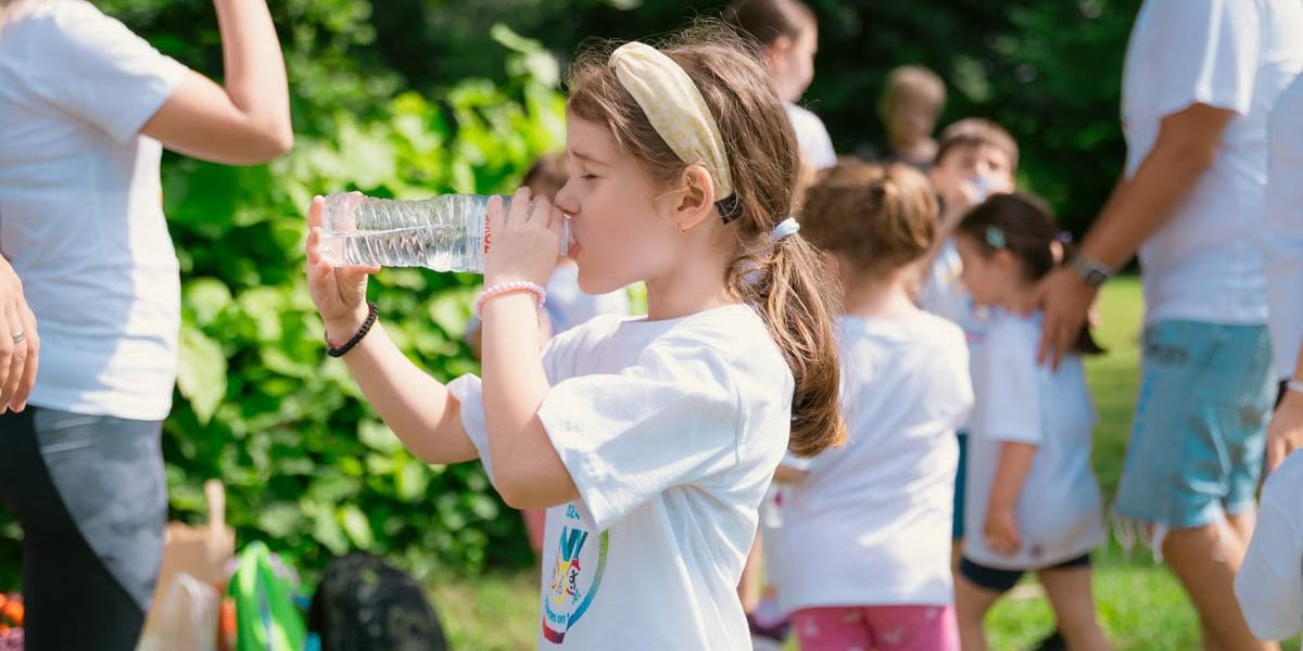 Young girl drinking water during sunny outdoor activity with friends in a park setting.