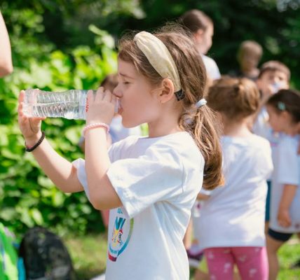 Young girl drinking water during sunny outdoor activity with friends in a park setting.